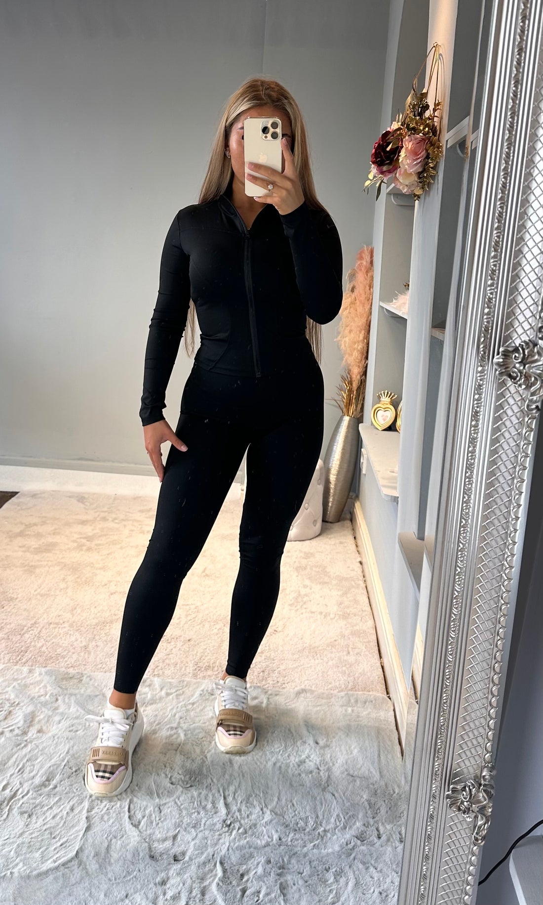Woman posing in a black full-zip fitted jumpsuit with long sleeves and sculpted leggings, ideal for workout, athleisure, or everyday wear, styled with beige sneakers in a modern interior setting.