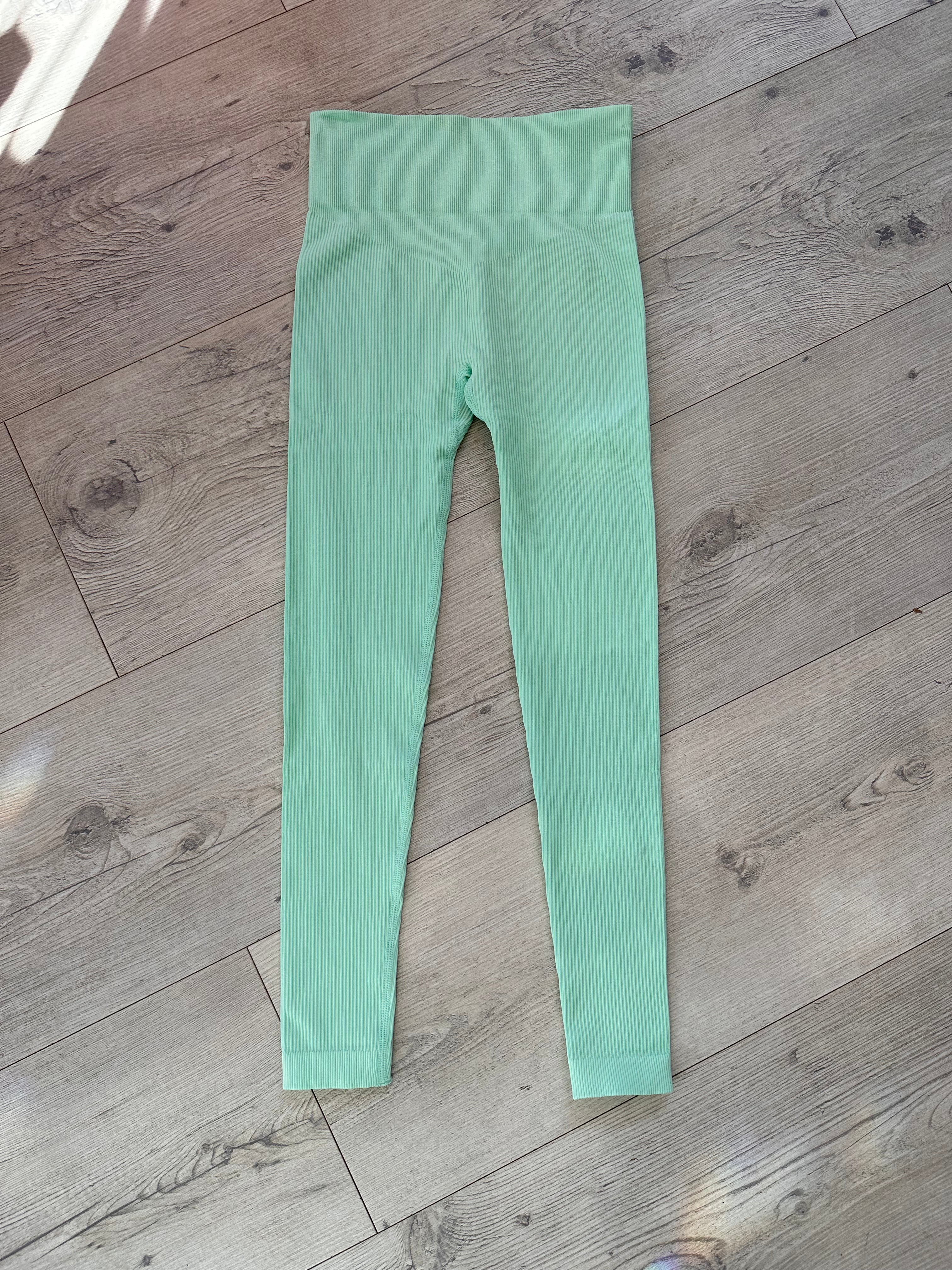 Flat lay of mint green ribbed leggings with a high-rise V-shaped waistband, displayed on a wooden floor with soft natural light