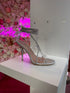 Silver clear strap stiletto heels for women with rhinestone wrap ankle detail, displayed on white pedestal with pink floral background
