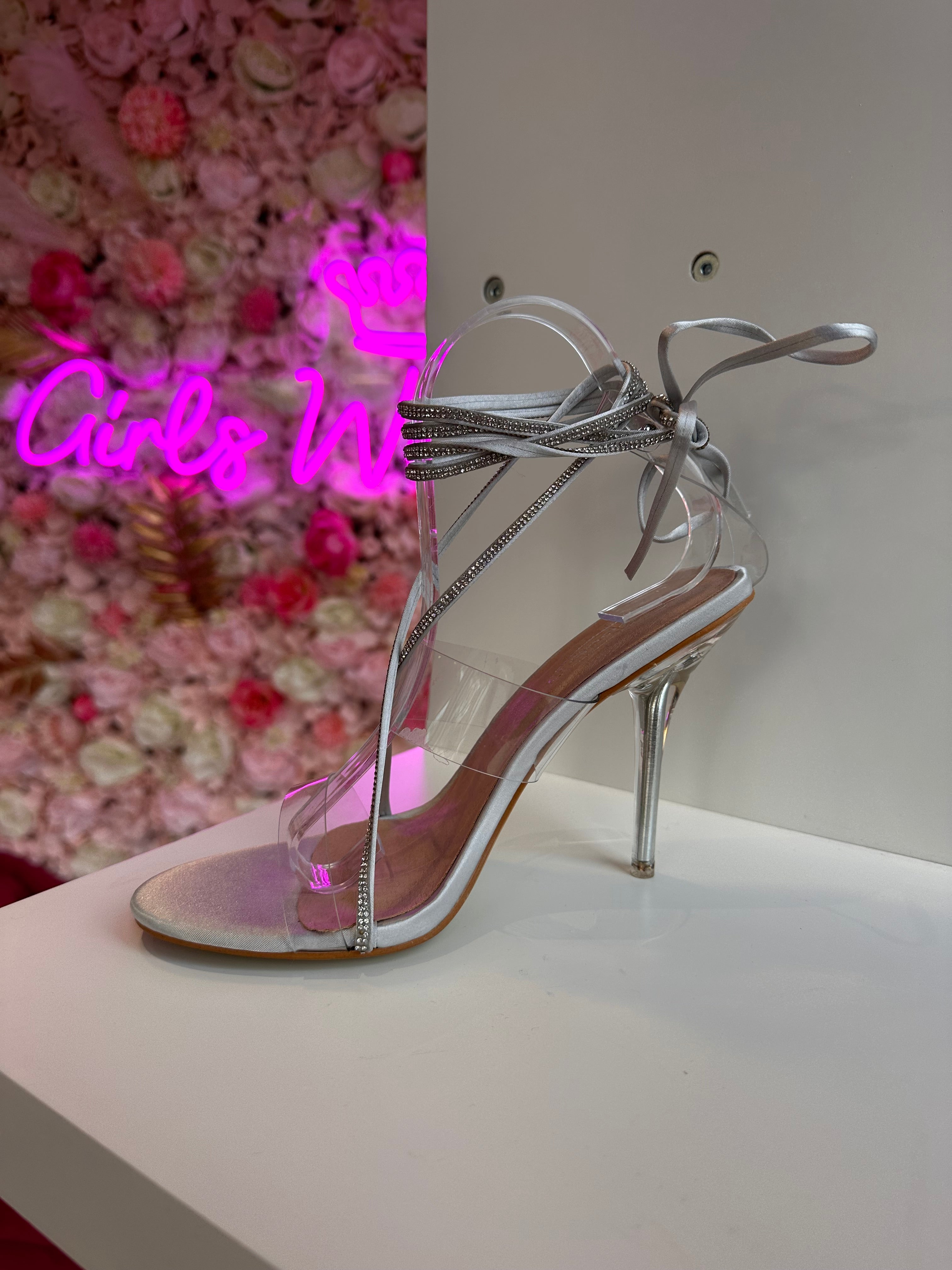 Stylish silver high heels with clear PVC straps and rhinestone lace-up ankle ties, displayed on a white shelf with floral pink backdrop