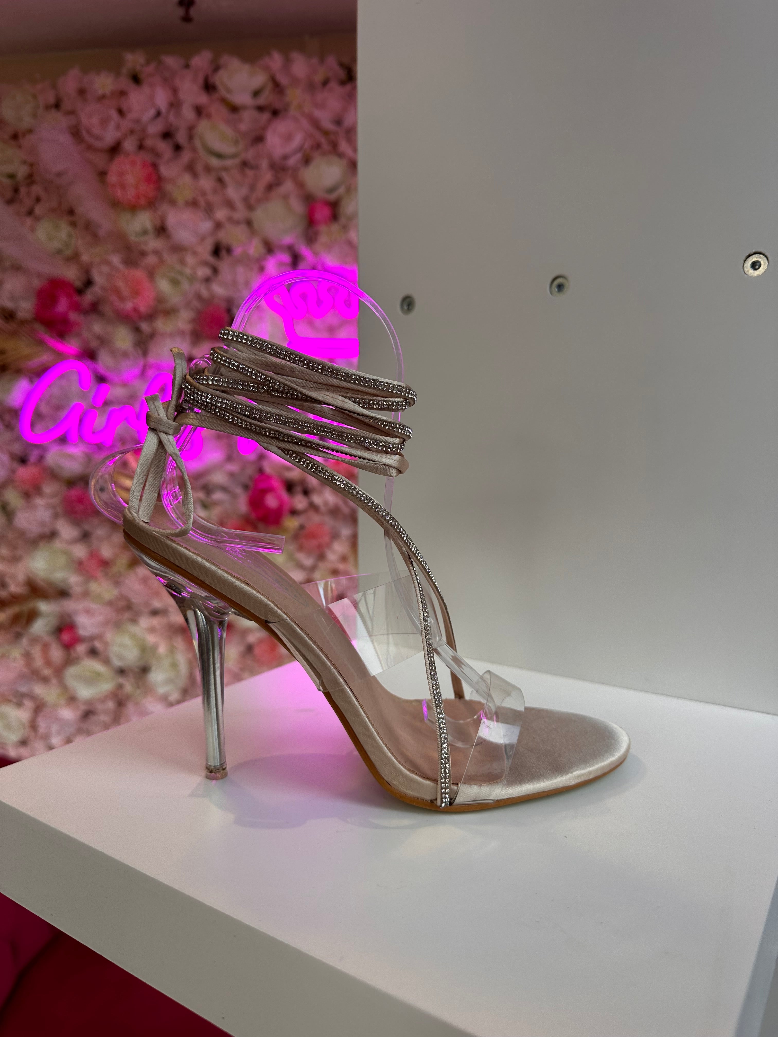 Chic transparent high heel sandal for women featuring rhinestone wrap straps and metallic stiletto heel, displayed on shelf with floral boutique background
