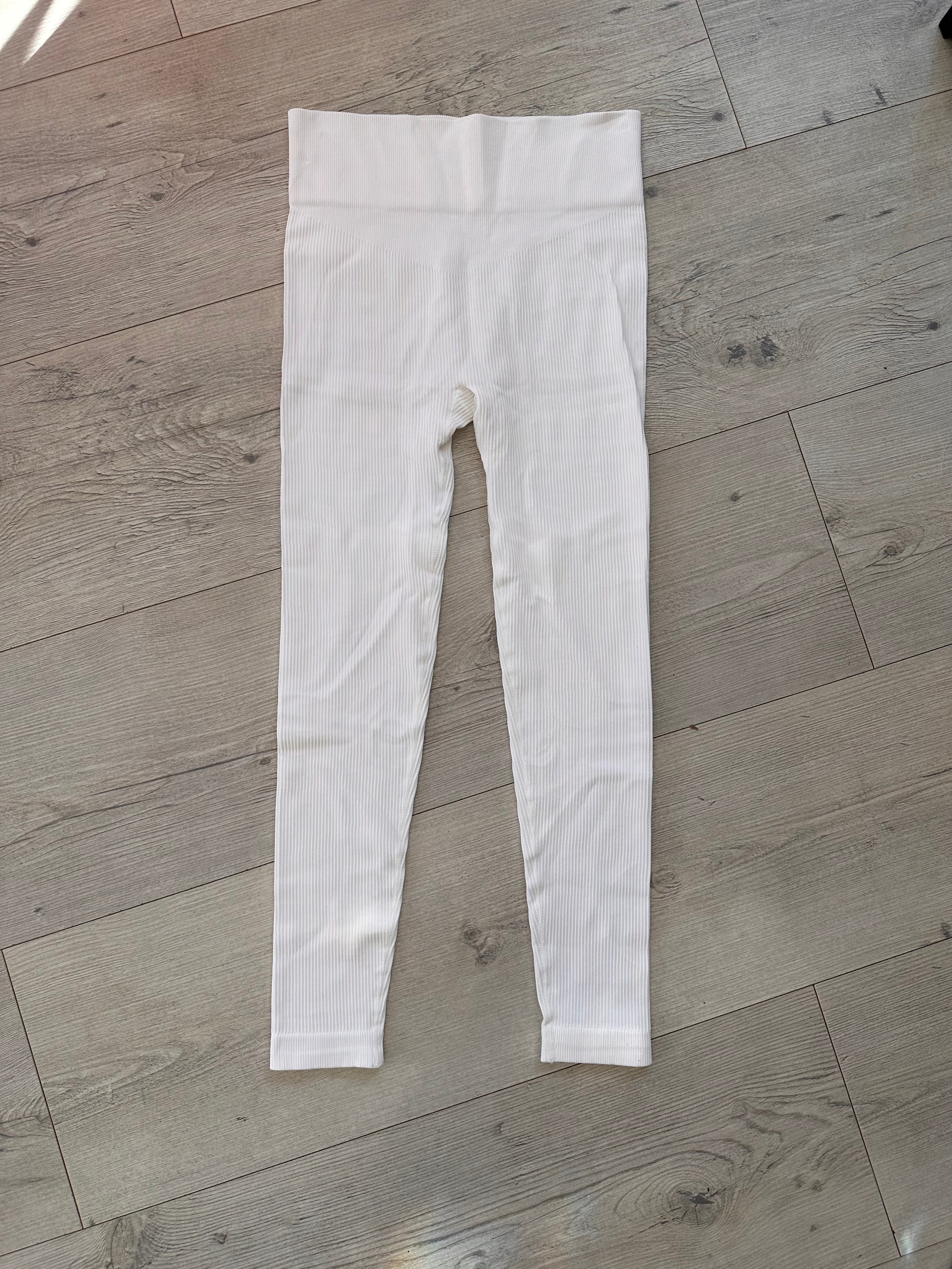 Flat lay of white ribbed leggings with a high-rise V-shaped waistband, displayed on a wooden floor under soft natural lighting