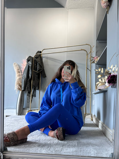 Woman wearing a bright blue ribbed leggings and oversized hoodie set, sitting on a soft rug and taking a mirror selfie in a modern room with neutral decor and gold clothing rack