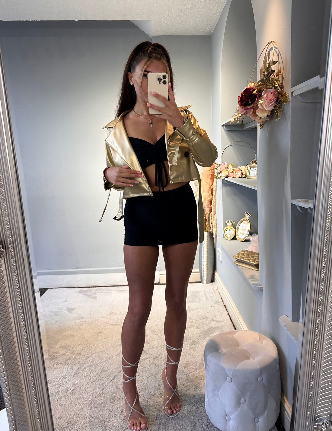 Woman wearing gold metallic trench coat over black mini dress and lace-up heels, posing in front of mirror for a chic night-out look – perfect party-ready outfit for women