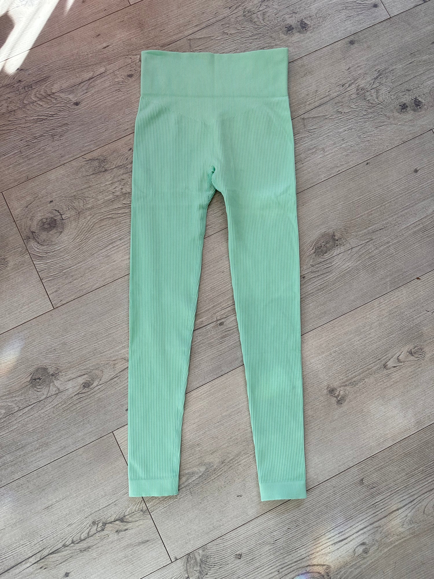 Flat lay of mint green ribbed leggings with a high-rise V-shaped waistband, displayed on a wooden floor with soft natural light