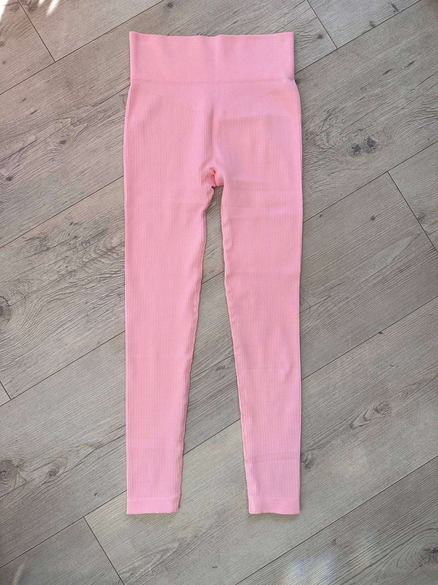 Flat lay of pink ribbed high-waisted leggings with a contour waistband, displayed on a light wood floor under natural sunlight