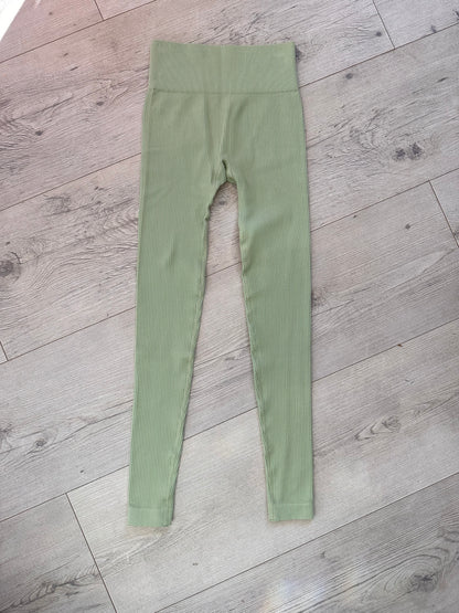 Flat lay of sage green ribbed leggings with a straight high-rise waistband, neatly displayed on a light wooden floor in natural lighting