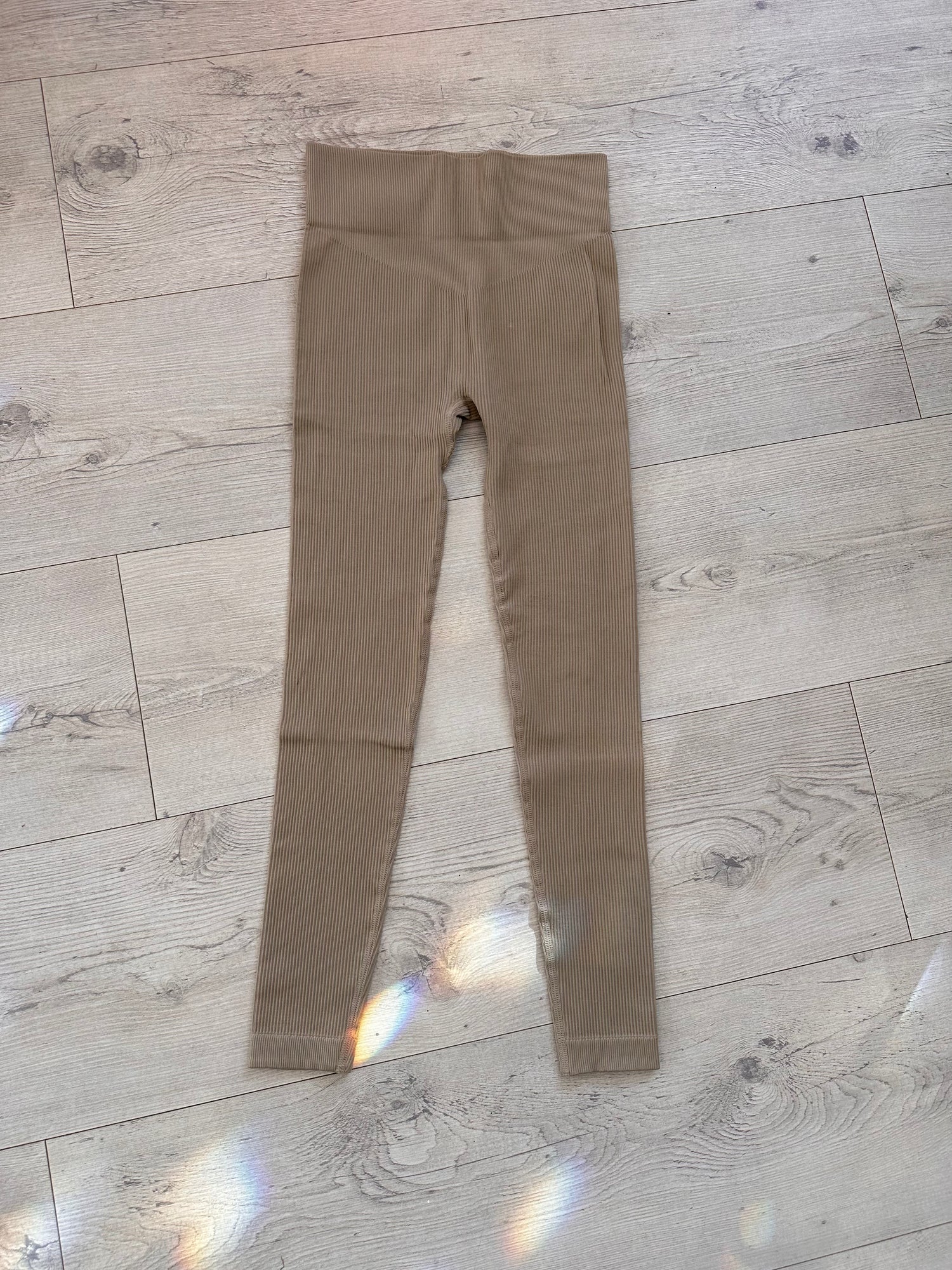 Flat lay of taupe ribbed high-waisted leggings with contour waistband, displayed on a light wood floor with soft rainbow light reflections